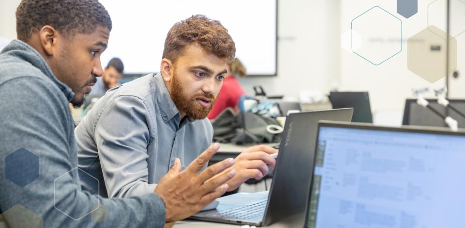 Two men sitting at a desk and looking at a laptop screen. 