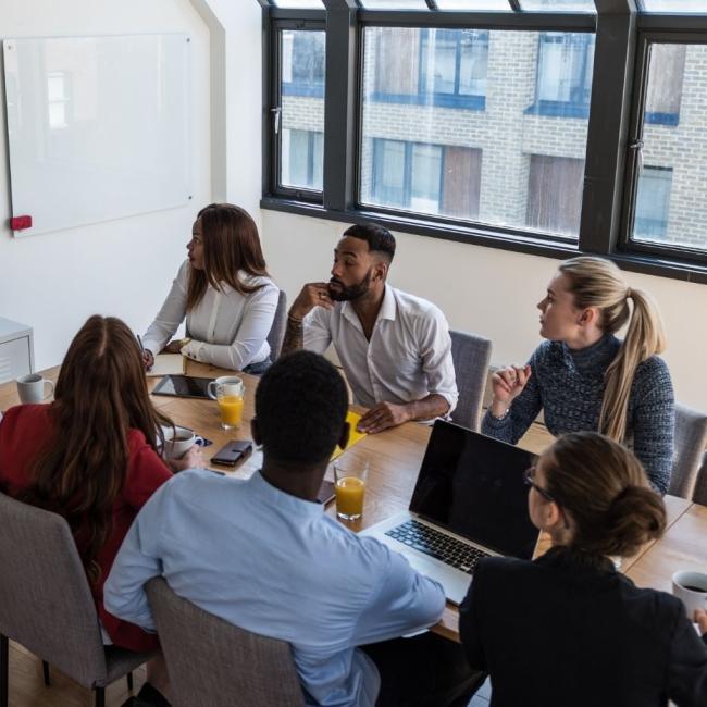 Working professionals conducting a hybrid business meeting, with some colleagues in a conference room and one colleague on video conference.